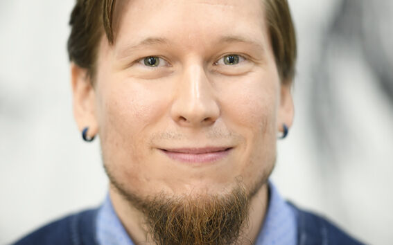 A man with a light brown beard, wearing a blue dotted shirt and a dark blue jumper.