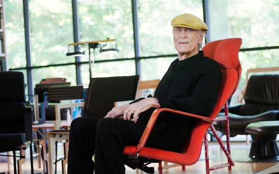 An elderly man wearing a yellow cap sitting in a red chair with black clothing in a bright, modern room with large windows.