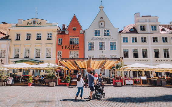 People walk in front of colourful historic buildings and outdoor restaurants in a sunny town square.