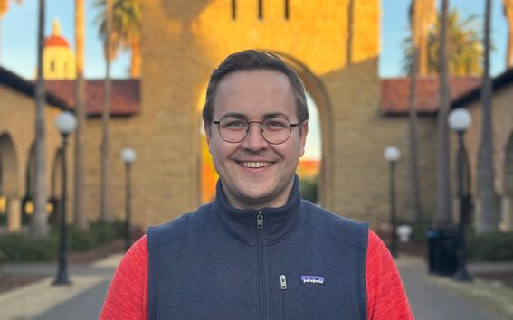 Person in a red-sleeved jacket standing on a pathway in front of a historic building with an arched entrance and palm trees.