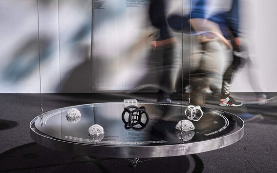 An art installation with geometric shapes on a round reflective table, people in motion are blurred in the background.
