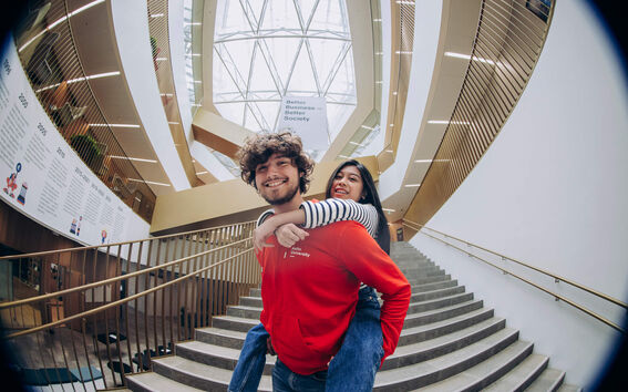 A person in a red hoodie giving another a piggyback ride in a modern building with a large glass ceiling and staircase.