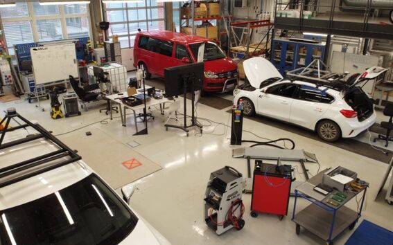 Työpaja, jossa kaksi autoa, punainen pakettiauto ja valkoinen hatchback. Näkyvillä laitteistoja ja osia.