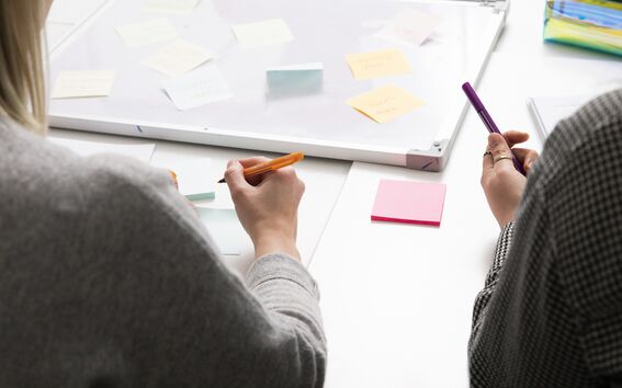 Kaksi henkilöä kirjoittaa pöydän ääressä värikkäillä kynillä muistilappuihin. Pöydällä makaa taulu, johon on kiinnitetty lisää muistilappuja.