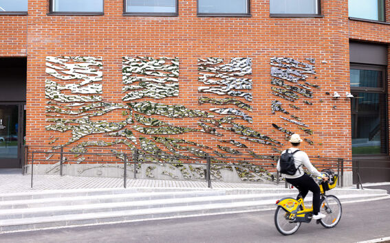 A person rides a yellow bike past a brick wall with abstract metal art installations. The rider wears a hat and has a backpack.