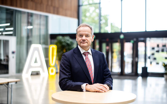 Henkilö puvussa ja punaisessa raidallisessa solmiossa seisoo pyöreän pöydän ääressä sisätiloissa, lähellä suurta hehkuvaa A!-merkkiä.