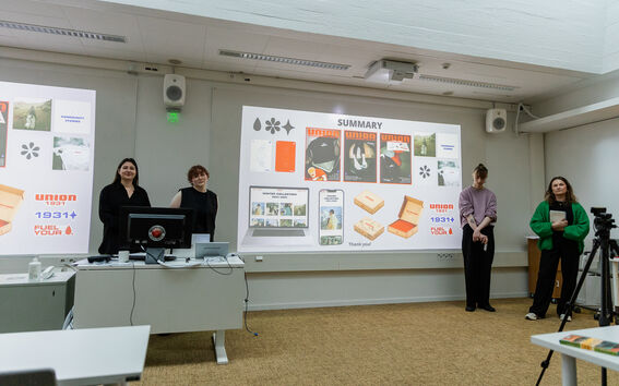 Presentaatio luokassa, jossa seisoo kolme opiskelijaa, kaksi näyttöä näyttää projektien yhteenvedot ja kuvat.