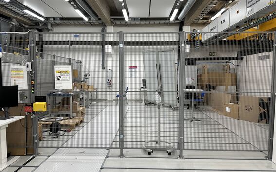 Varasto, jossa metalliaita, pahvilaatikoita, pöydät varusteineen ja varoitusmerkki. Kirkkaat kattovalot valaisevat aluetta.