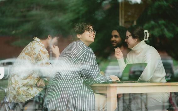 Ryhmä ihmisiä keskustelemassa puisen pöydän ääressä kahvilassa. Pöydällä näkyy yksi kannettava tietokone.