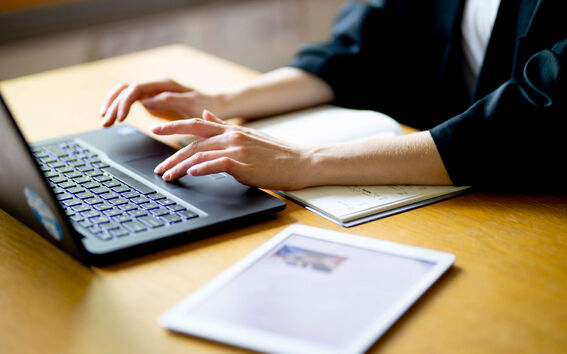 A person typing on a laptop next to a notebook and tablet on a wooden desk.