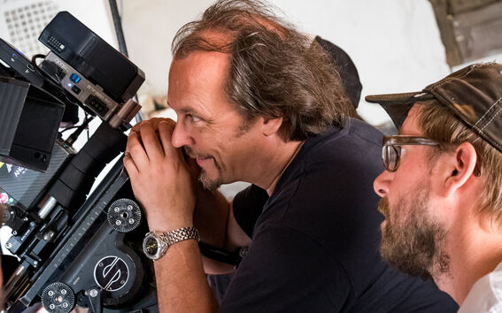 Kaksi ihmistä käyttää ammattilaistason elokuvakameraa. Toinen pukeutunut mustaan paitaan, toinen lippalakkiin.