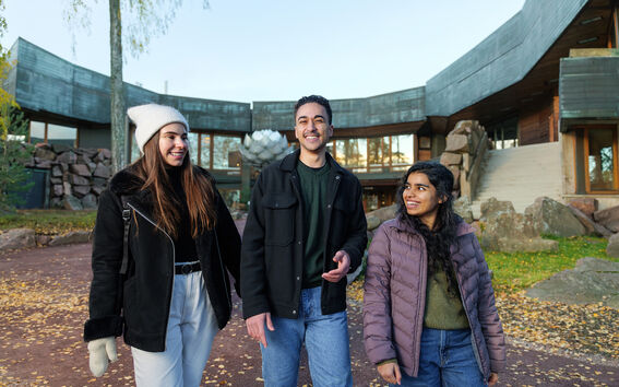 Kolme ihmistä kävelemässä ulkona modernin rakennuksen lähellä, jossa on suuret lasi-ikkunat ja kiviseinät.