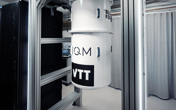 A white cylindrical quantum computer with the logos IQM and VTT, installed in a framework in a laboratory.