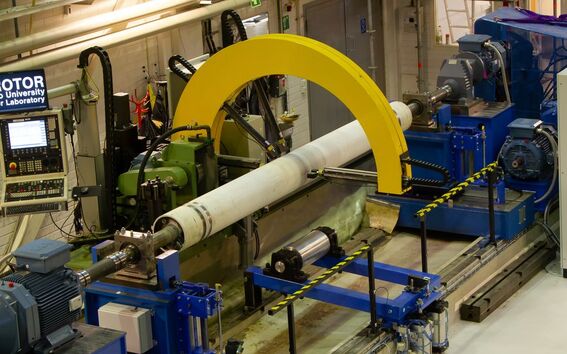 Teollinen roottorin testauskone, jossa keltainen kaari, siniset komponentit ja ohjauspaneelit Aalto-yliopiston roottorilaboratoriossa.