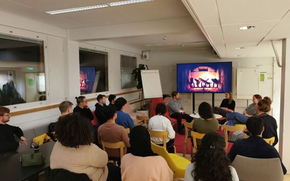 A group of people gathered in a modern indoor setting, attentively listening to a panel discussion. Two guest speakers are seated in front of the audience, engaged in conversation. A large screen behind them displays a digital fireplace, creating a warm atmosphere. The attendees sit on chairs arranged in a semi-circle, actively participating in the discussion.