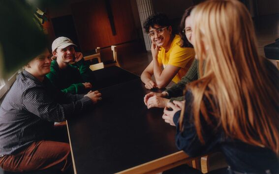 Kuusi henkilöä istuu tumman pöydän ääressä modernissa, avoimessa tilassa. Yhdellä on pitkät hiukset ja tummat vaatteet.