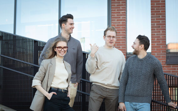 Neljä henkilöä rennossa vaatetuksessa seisoo portailla rakennuksen ulkopuolella, jossa on suuret ikkunat ja tiiliseinät.