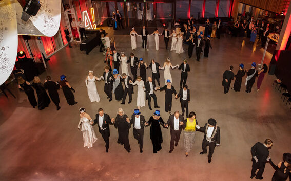 En formell dans, där människor klädda i festkläder håller händer i en stor cirkel. Några bär blå hattar.