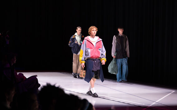 Kolme mallia kävelee catwalkilla. Etummainen malli pukeutunut värikkääseen takkiin, mustiin shortseihin ja mustiin kenkiin.