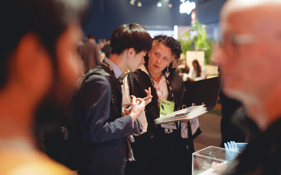 Kaksi ihmistä keskustelee sisätapahtumassa. Toinen pitää kansiota, ja toinen viittoo kädellään.