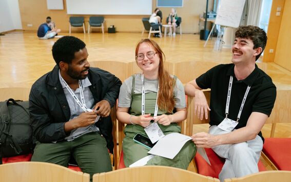 Kolme ihmistä istumassa sisätiloissa tuoleilla, pitävät papereita kädessä ja juttelevat. Taustalla istuu ihmisiä ryhmissä.