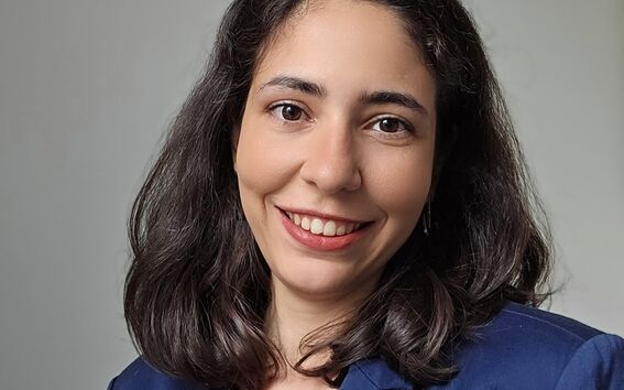 A woman with shoulder-length dark hair wearing a blue blazer and a white top. Grey background.
