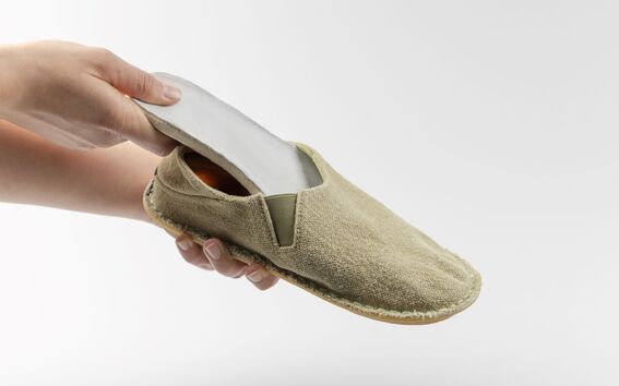 A hand holding a beige slip-on shoe, removing the insole from the inside. The background is plain white.