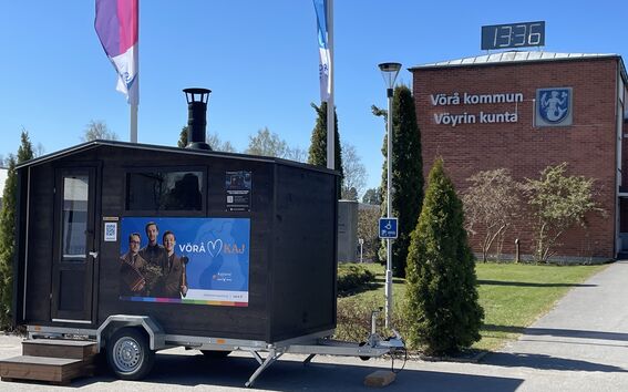 Liikuteltava sauna ja Vöyrin kunnanvirasto taustalla. Kolme lippua ja suuri digitaalinen kello näkyvissä.