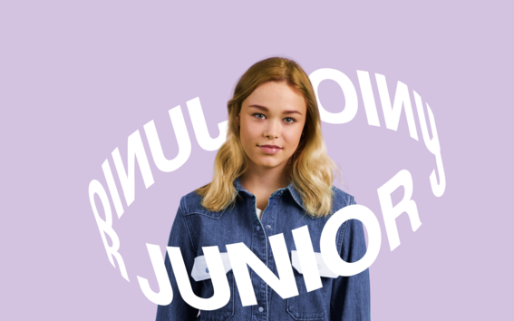 Henkilö, jolla on farkkupaita ja vaaleansiniset farkut seisoo vaalean violetilla taustalla. Teksti 'JUNIOR'.