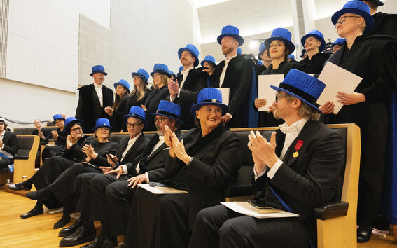 People wearing black academic robes and blue caps in a ceremony. Some are standing, others clapping from seats.