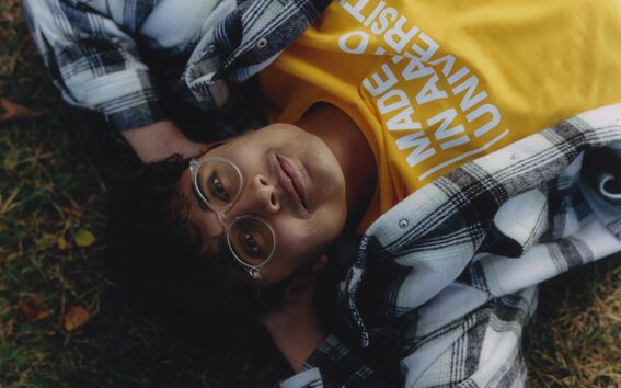 Person wearing a yellow shirt with text 'Made in Aalto University' and a black-and-white plaid jacket, lying on grass.