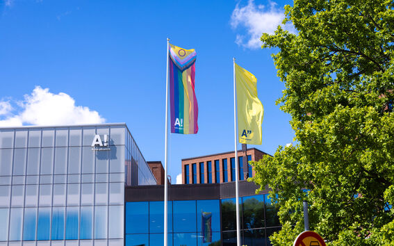 Aalto-yliopistolla kaksi lippua: sateenkaarilippu ja keltainen lippu. Taustalla moderni rakennus ja vihreitä puita.