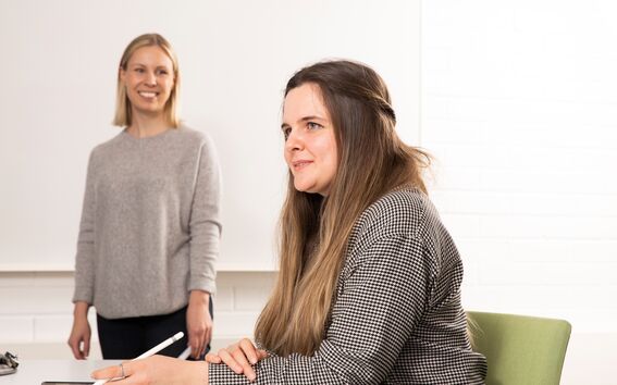 Kaksi hymyilevää henkilöä, joista toinen kuuntelee seisten ja toinen istuu kynä kädessä ja puhuu.