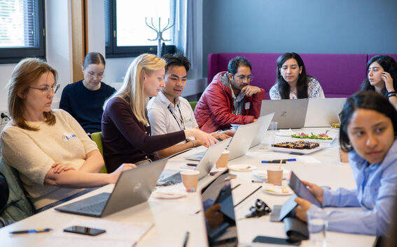 A group of people sitting at a table with laptops and snacks, engaged in a discussion in a modern office setting.
