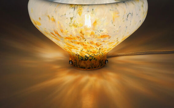 Koristeellinen lasilamppu, joka tuottaa lämpimän hehkun, asetettuna tummalle pinnalle. Sen valo loistaa kuvioidun lasikulhon läpi.