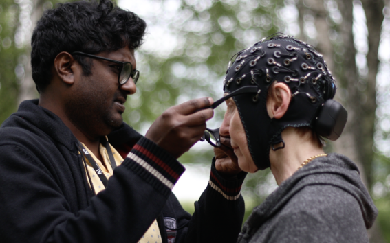 In a park-like setting, a researcher helps a participant put on a mobile imaging device. The device captures data such as eye movements and pupil reactions to assess risks related to memory functions.