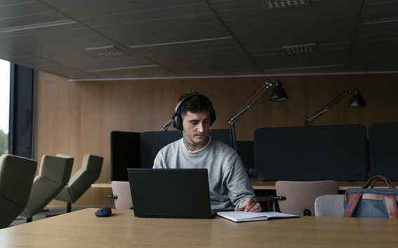 Henkilö kuulokkeissa työskentelee kannettavalla tietokoneella pöydän ääressä, taustalla puinen seinä ja suuret ikkunat.