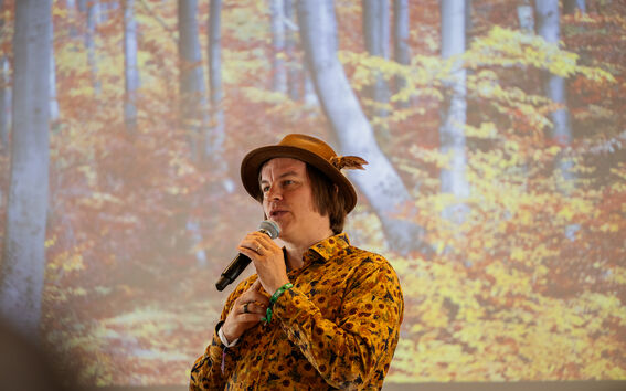 A person wearing a colourful shirt and brown hat speaks into a microphone with a background of an image of a forest.