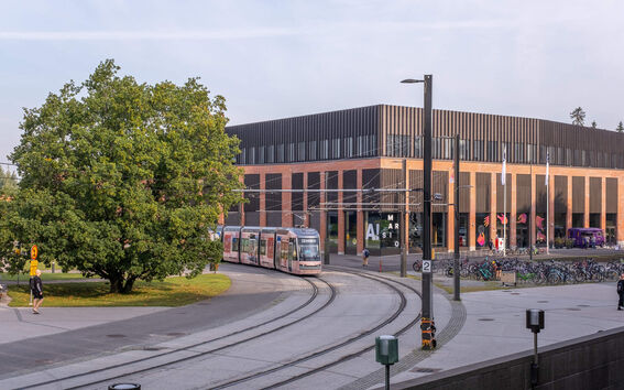 Moderni raitiovaunu kulkee puun ja tiilirakennuksen ohitse, jossa on suuret ikkunat.