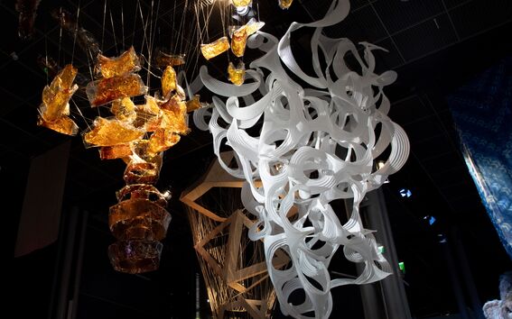 Large artworks hanging on ceiling in a dark room: amber-like resin sculpture and swirly white paper sculpture.