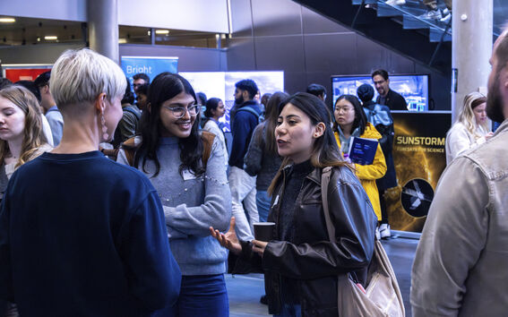 Ryhmä ihmisiä vilkkaassa sisätilaisuudessa näyttelyosastoilla. Yksi henkilö pitelee kuppia ja puhuu elehtien.