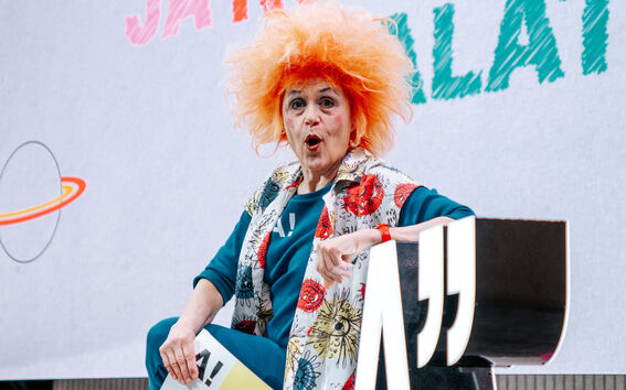 Person with orange hair sits on stage holding a sign with '15', near large letters 'A' and quotation marks.