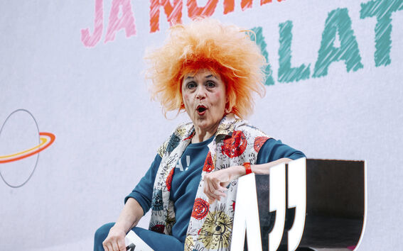 Oranssihiuksinen henkilö istuu lavalla ison A-kirjaimen vieressä ja pitelee kylttiä, jossa on numero 15.