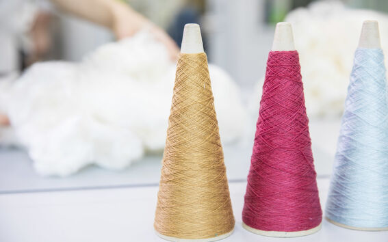 Three cones of thread in gold, pink, and light blue with a blurred background of white fabric and a hand.