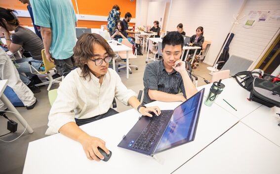 Kaksi ihmistä yhteisellä pöydällä käyttämässä kannettavaa tietokonetta, ympärillä muita työskentelemässä modernissa, kirkkaanvärisessä huoneessa.