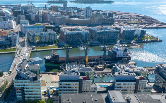 Aerial view of modern urban buildings with rooftop gardens by the sea. A ship and cranes are visible in the harbour.