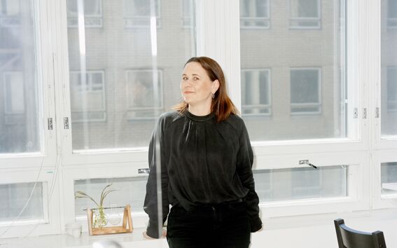 Henkilö seisoo sisätiloissa valkoisen tason ääressä korkeiden ikkunoiden edessä, yllään mustat vaatteet ja kengät.