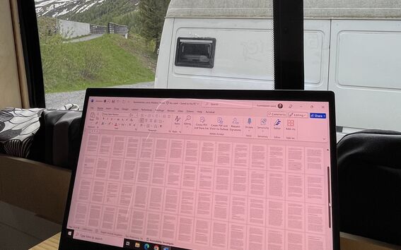 A laptop with a document displayed sits on a wooden table inside a camper van, with a snowy mountain visible through the window.