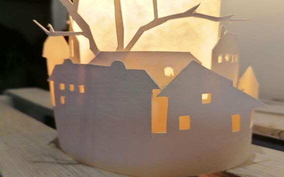 Paperilyhty kuvaa taloja ja puita leikattuina kuvioina, lämmin valo valaisee sisältä.