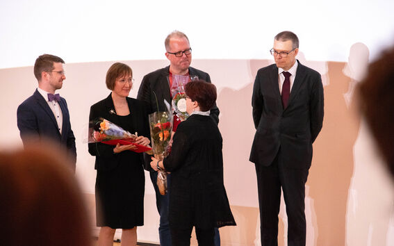 Four awarded individuals dressed in dark clothing receiving bouquets at the academic year's opening ceremony.